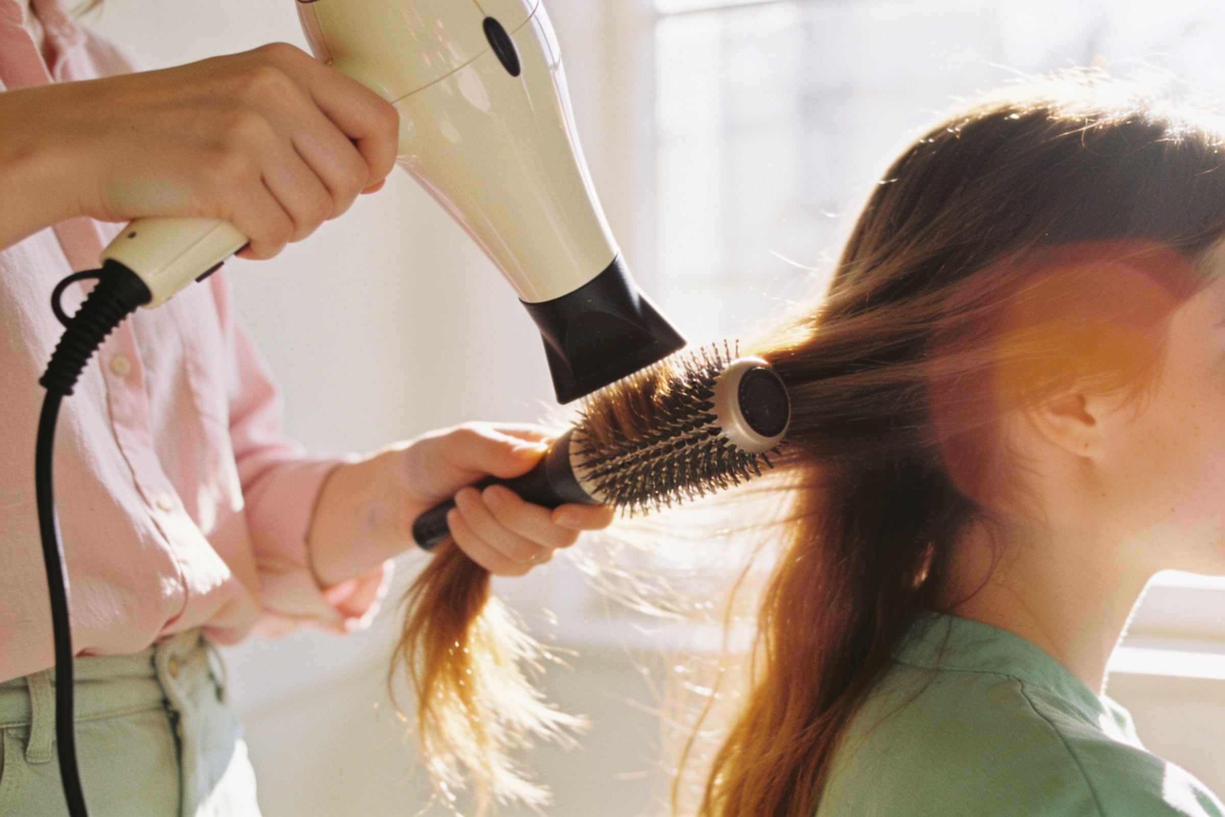 A stylist blow-drying long glossy hair with a round brush in warm sunlight.