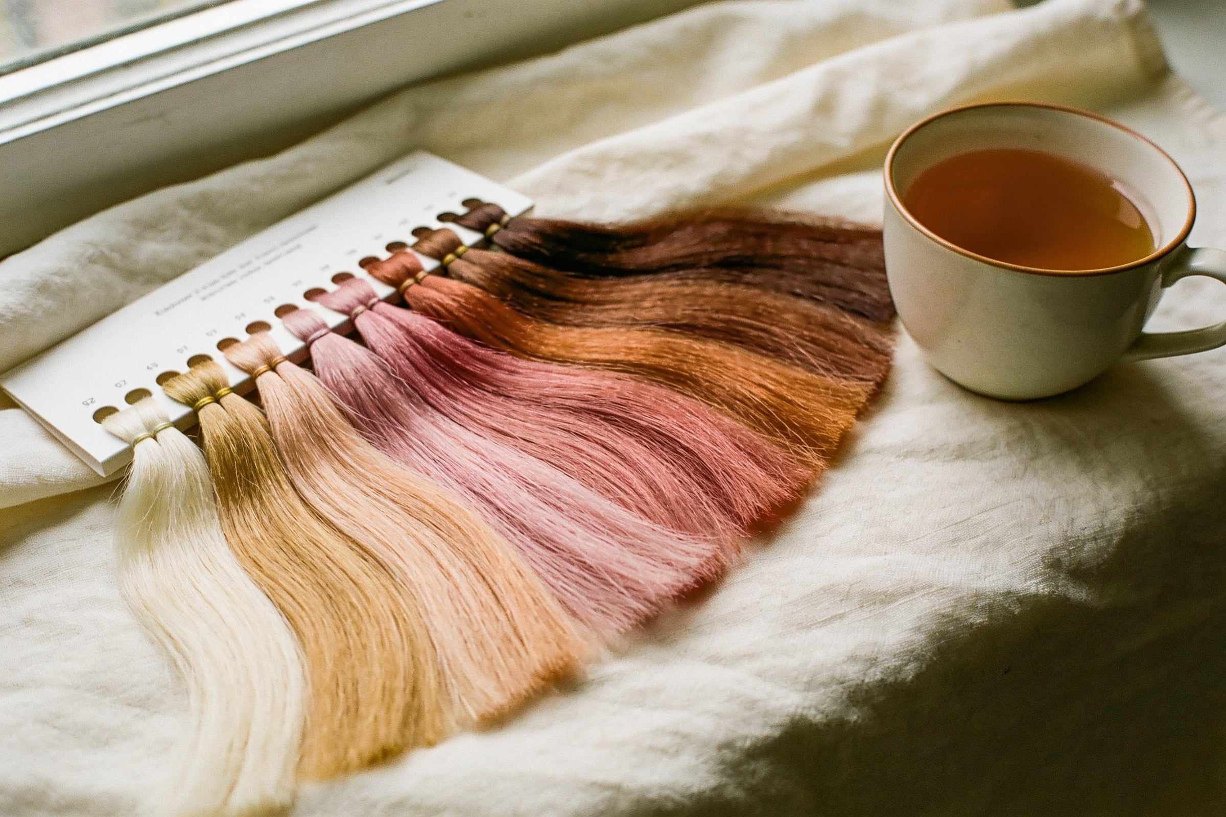A fan of hair color swatches from pale blonde through honey, rose and copper to chestnut, beside a cup of tea.