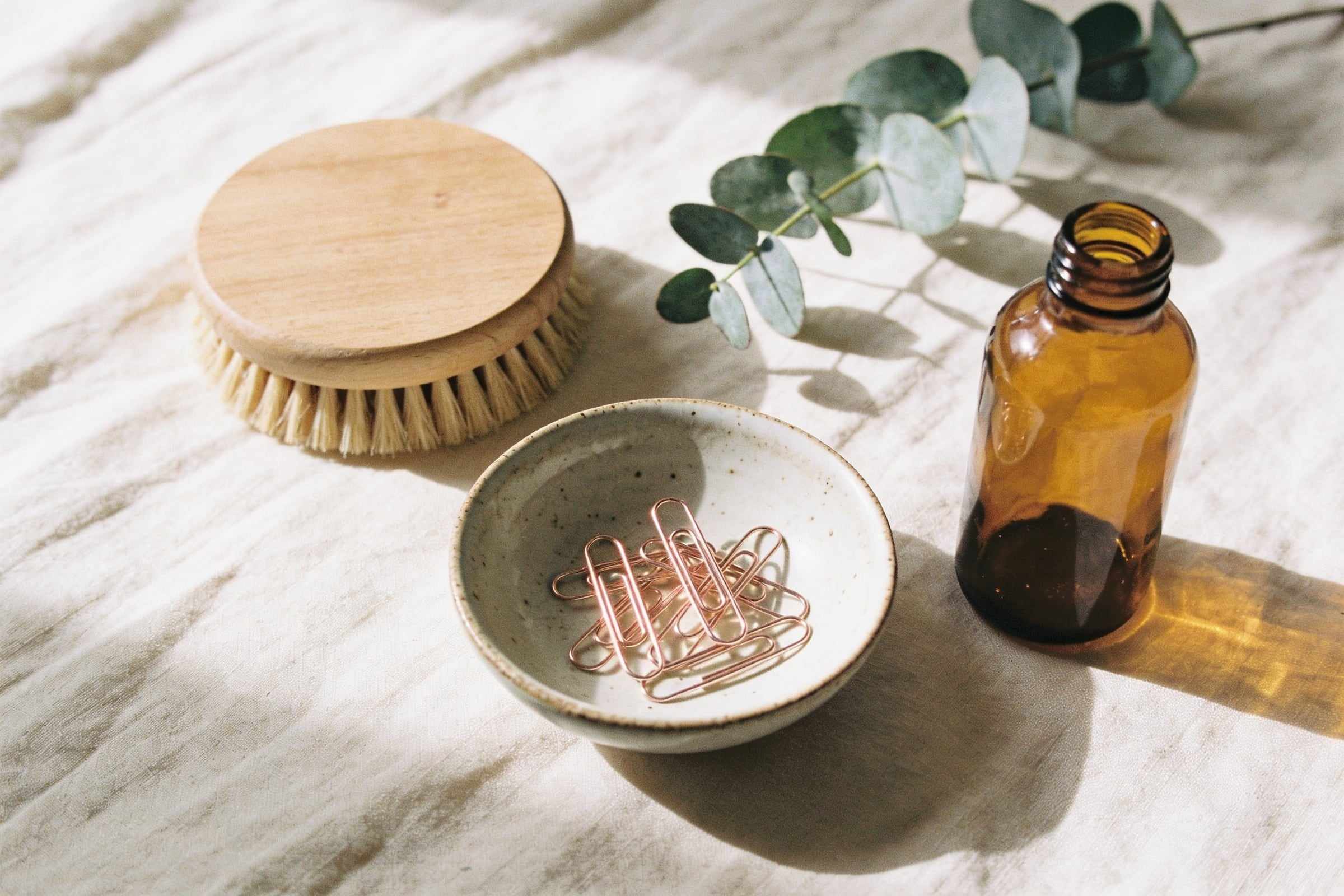 A wooden round brush, hair clips in a ceramic dish and a sprig of eucalyptus on cream linen.