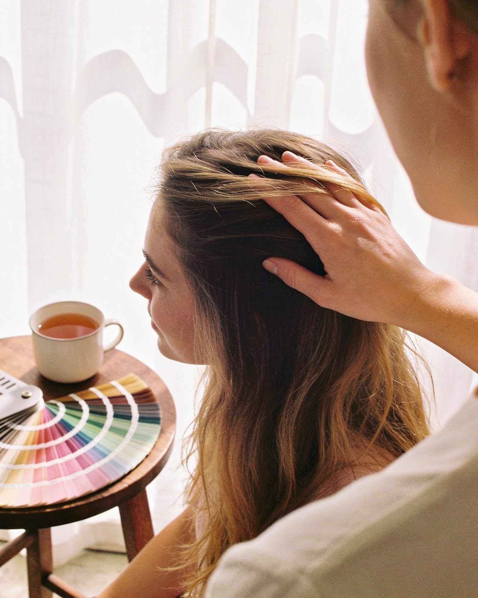 A stylist rests a hand on a client’s hair beside a cup of tea and a ring of color swatches.