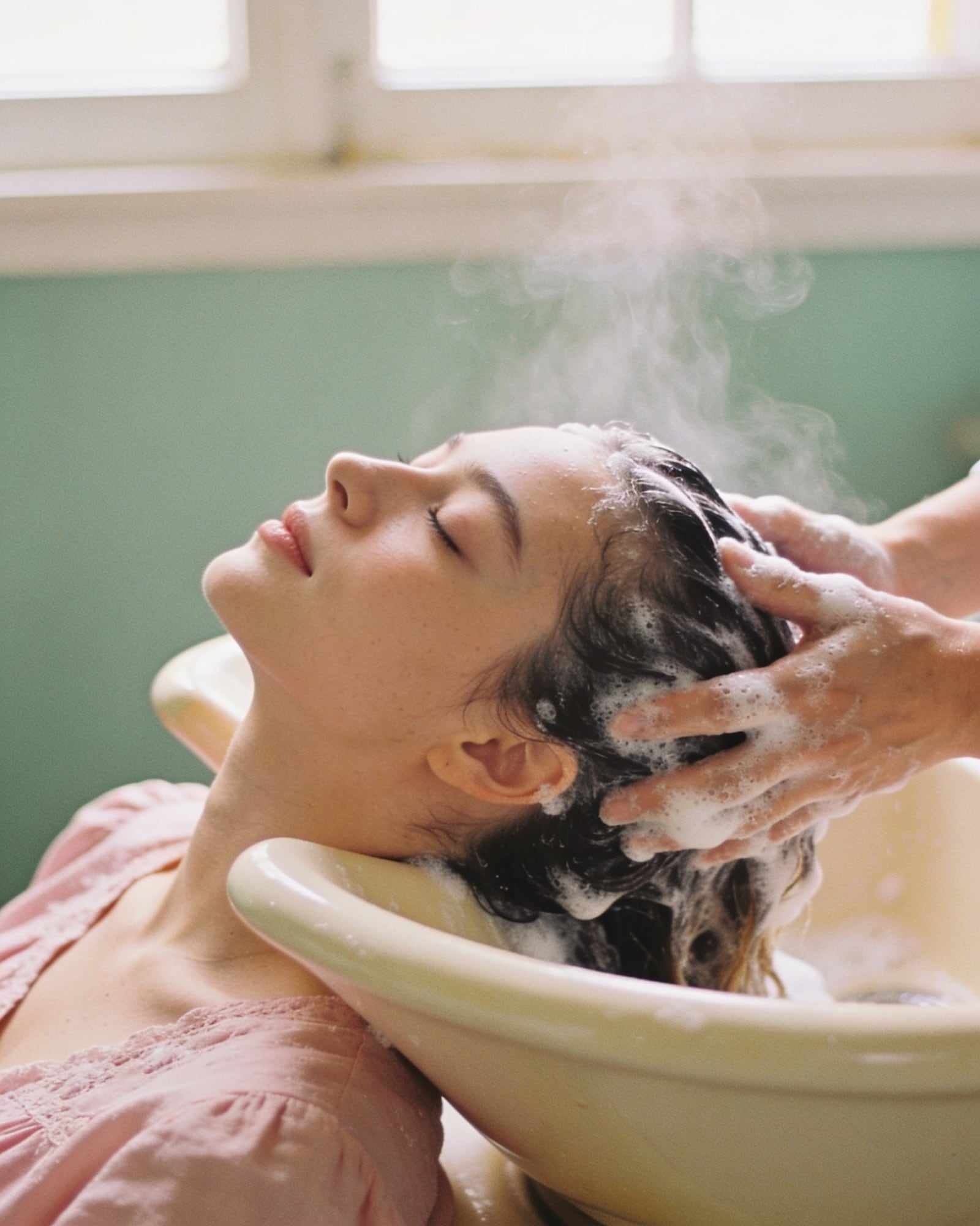 A woman leans back at a cream basin, eyes closed, while her hair is washed.